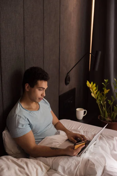 Schöner gemischter Rassenmann beim Online-Shopping mit Kreditkarte und Laptop im Bett unter Quarantäne — Stockfoto