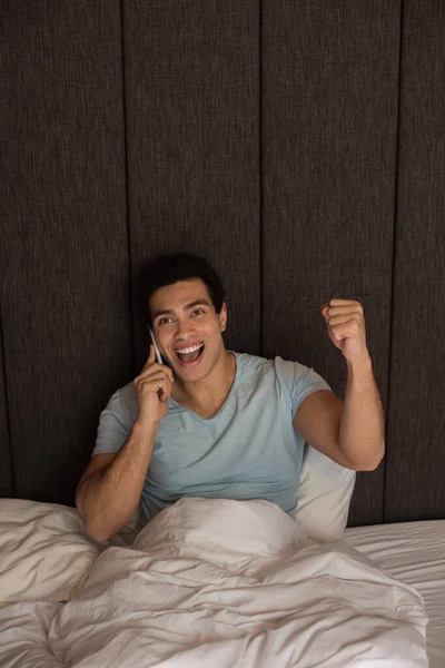 Взволнованный смешанной расы человек говорит на смартфоне в постели во время самоизоляции — стоковое фото