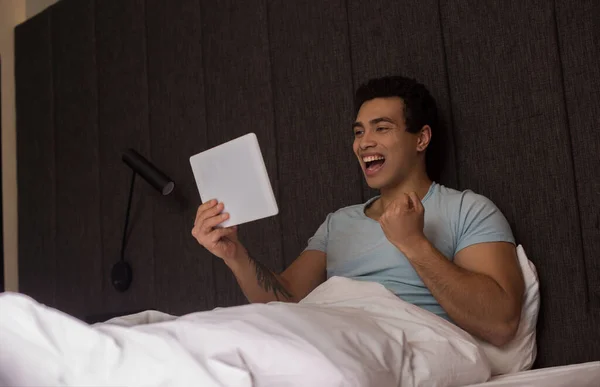 Excité beau mixte homme utilisant tablette numérique au lit pendant la quarantaine — Photo de stock