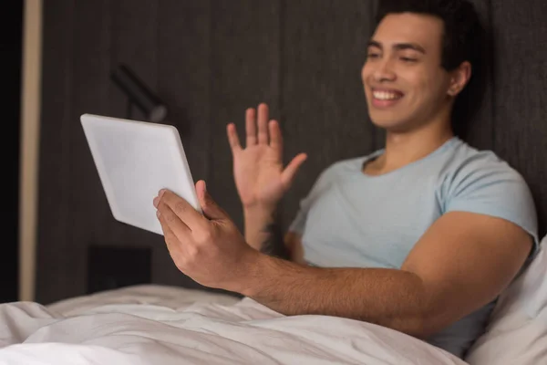 Glücklicher Mann mit gemischter Rasse winkt, während er während der Selbstisolierung einen Videochat auf einem digitalen Tablet im Bett führt — Stockfoto