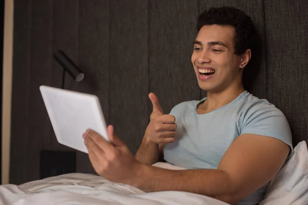 Guapo feliz mixto hombre mostrando el pulgar hacia arriba mientras tiene videollamada en tableta digital en la cama durante la cuarentena - foto de stock