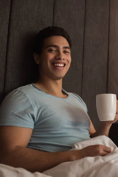 Sonriente hombre de raza mixta sosteniendo taza de café en la cama en cuarentena - foto de stock