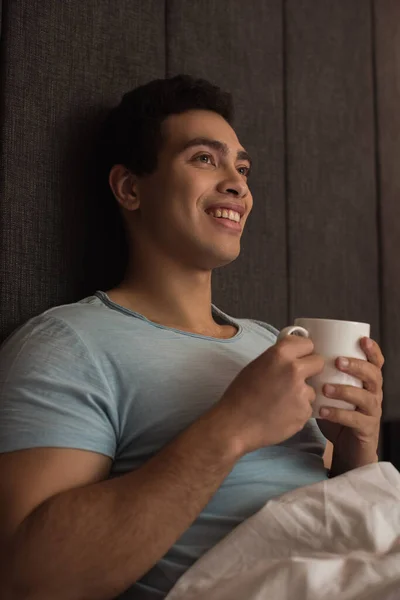 Sorrindo misto raça homem segurando xícara de café na cama em auto-isolamento — Fotografia de Stock