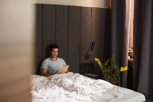 Happy mixed race man reading book in bed during self isolation, selective focus — Stock Photo