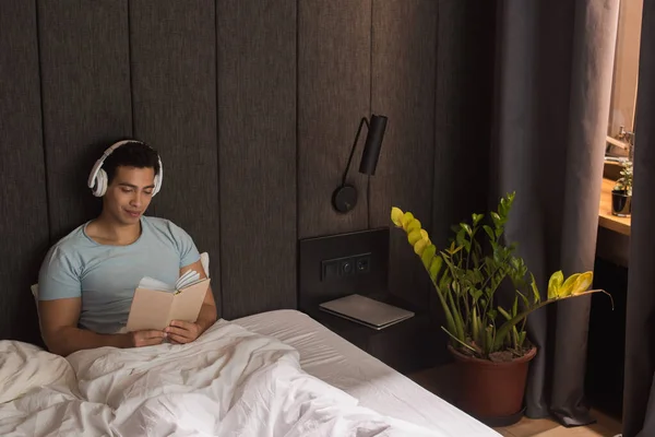 Happy mixed race man reading book while listening music with headphones in bed during self isolation — Stock Photo