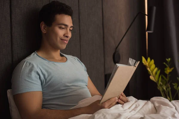 Sonriente mestizo hombre leyendo libro en la cama durante auto aislamiento - foto de stock