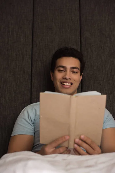 Smiling mixed race man reading book in bed during quarantine — Stock Photo