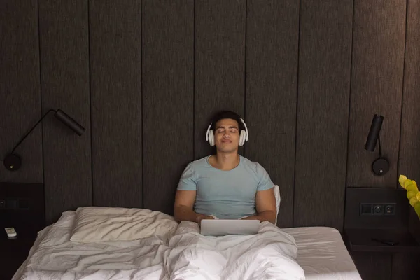 Hombre bi-racial con los ojos cerrados escuchando música con auriculares y portátil mientras se relaja en la cama en cuarentena - foto de stock