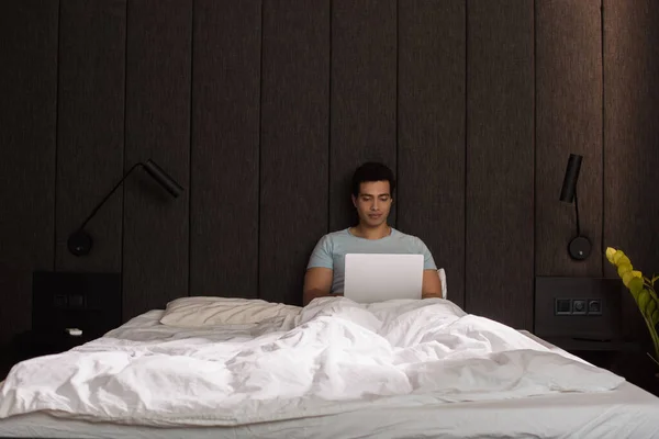 Freelancer masculino de raza mixta que trabaja en el portátil en la cama durante el autoaislamiento - foto de stock