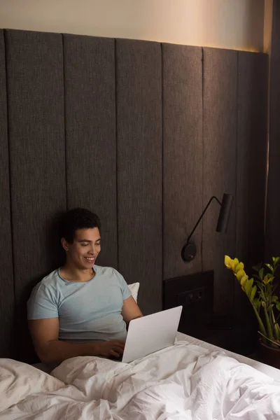 Happy male mixed race freelancer working on laptop in bed during self isolation — Stock Photo