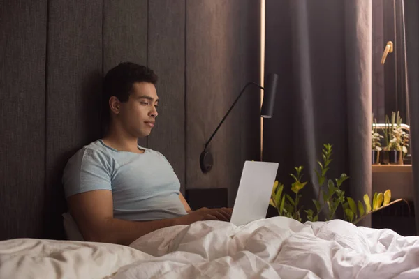 Beau mâle mélange race pigiste travaillant sur ordinateur portable au lit en quarantaine — Photo de stock