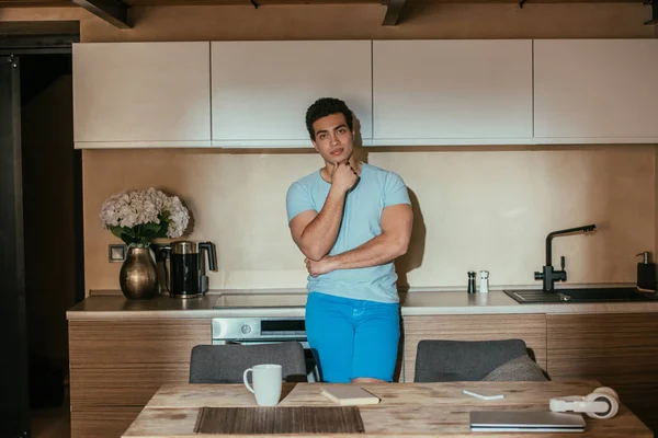 Pensativo hombre de raza mixta de pie en la cocina con taza de café, libro, auriculares y portátil durante la cuarentena - foto de stock