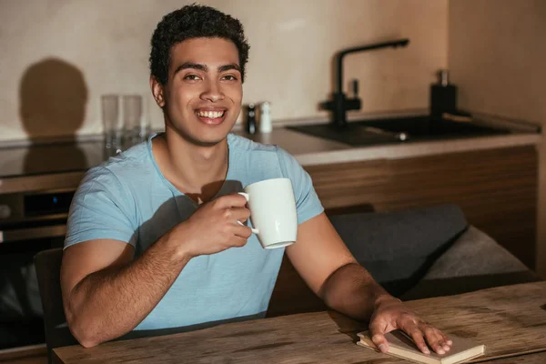 Sonriente hombre de raza mixta sosteniendo taza de café y libro en la cocina durante el auto aislamiento - foto de stock