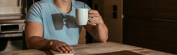 Vue recadrée de l'homme tenant une tasse de café et réservant sur la cuisine pendant l'isolement, orientation panoramique — Photo de stock