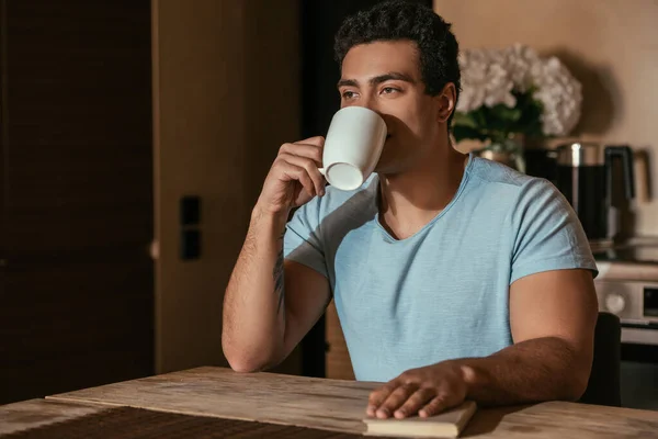 Mestiço homem bebendo café e segurando livro na cozinha durante o auto-isolamento — Fotografia de Stock