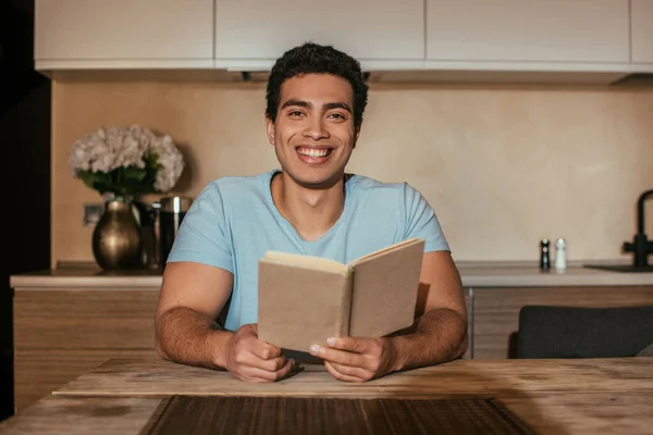 Sonriente mestizo hombre leyendo libro en cocina durante cuarentena - foto de stock