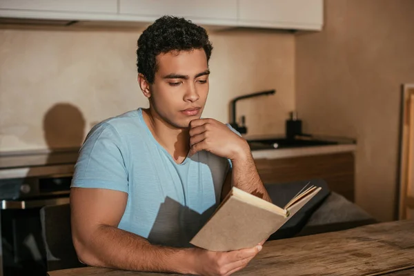 Pensativo bi-racial homem ler livro na cozinha durante a quarentena — Fotografia de Stock