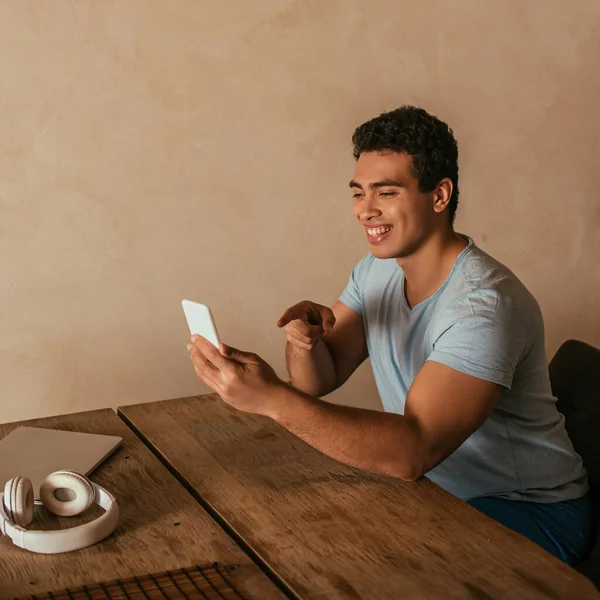 Feliz hombre de raza mixta señalando y tener videollamada en el teléfono inteligente en casa en cuarentena - foto de stock