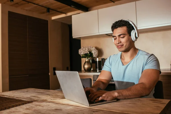 Happy mixed race man listening music with headphones and using laptop during self isolation on kitchen — Stock Photo