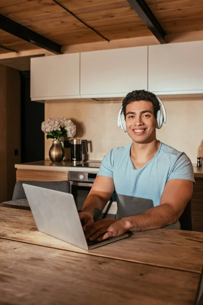 Homme de race mixte souriant écoutant de la musique avec des écouteurs et utilisant un ordinateur portable pendant l'isolement personnel sur la cuisine — Photo de stock