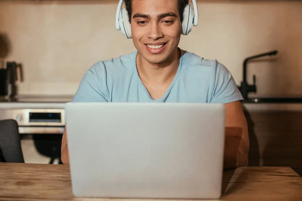 Hombre sonriente guapo escuchar música con auriculares y el uso de ordenador portátil durante el autoaislamiento en la cocina - foto de stock