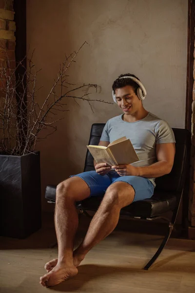 Счастливый смешанный человек расы читать книгу, слушая музыку с наушниками дома во время карантина — стоковое фото