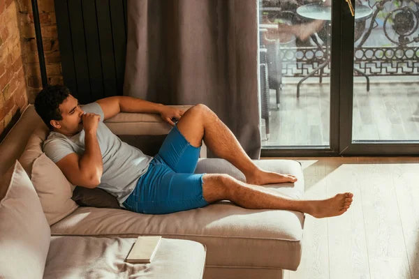 Homem de raça mista relaxando no sofá com livro na sala de estar durante a quarentena — Fotografia de Stock