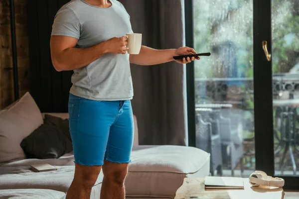 Vista recortada del hombre con taza de café sosteniendo mando a distancia y viendo durante el auto aislamiento - foto de stock