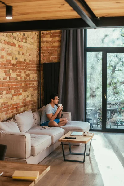 Красивий змішаний гоночний чоловік тримає чашку кави на дивані у вітальні під час карантину — стокове фото