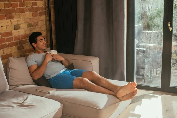 Alegre hombre de raza mixta sosteniendo taza de café en el sofá con libro durante la cuarentena - foto de stock