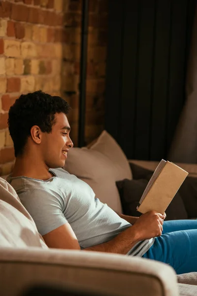 Bonito sorrindo misto raça homem leitura livro no sofá durante quarentena — Fotografia de Stock