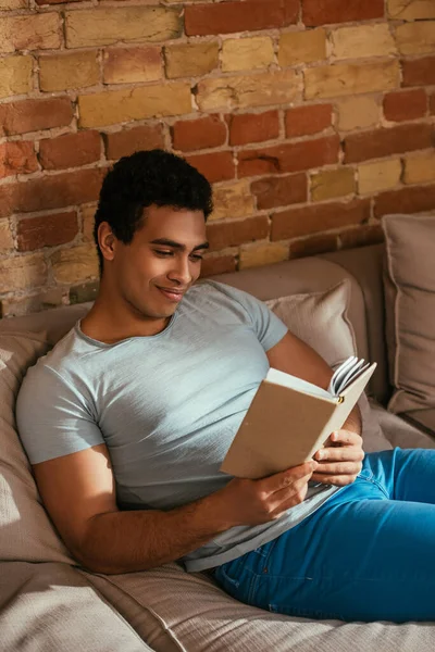 Homem de raça mista feliz ler livro no sofá durante a quarentena — Fotografia de Stock