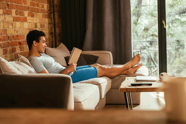 Lächelnder Mann mit gemischter Rasse liest während Quarantäne im Wohnzimmer Buch — Stockfoto