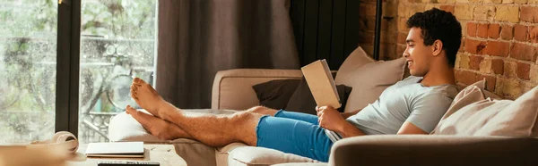 Smiling mixed race man reading book on sofa during quarantine, website header — Stock Photo