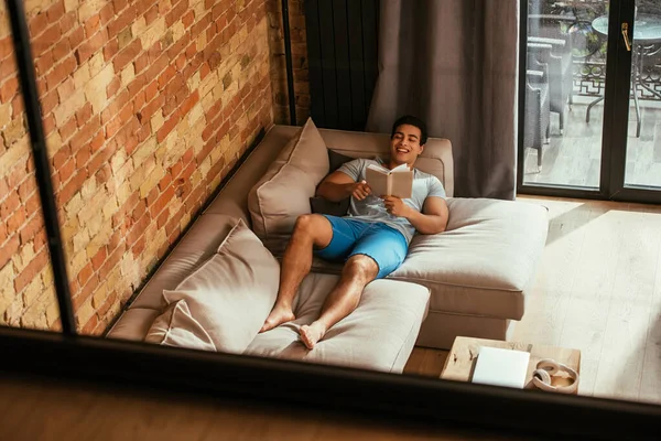 Смешанная раса человек читает книгу во время охлаждения на диване во время карантина — стоковое фото