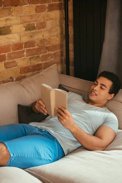 Красивый смешанный расовый человек читает книгу, расслабляясь на диване во время самоизоляции — стоковое фото