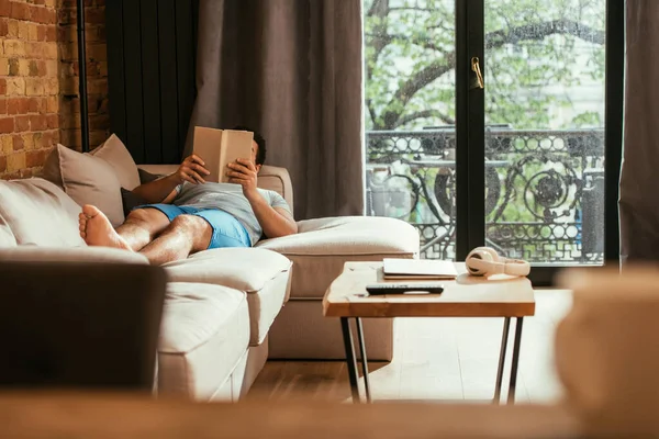 Joven leyendo libro mientras está acostado en el sofá durante la cuarentena - foto de stock