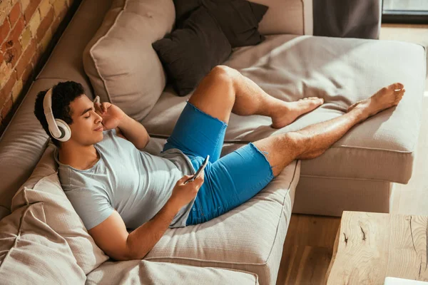 Schöner gemischter Rassenmann, der mit geschlossenen Augen chillt und während der Isolation Musik mit Kopfhörern und Smartphone auf dem Sofa hört — Stockfoto