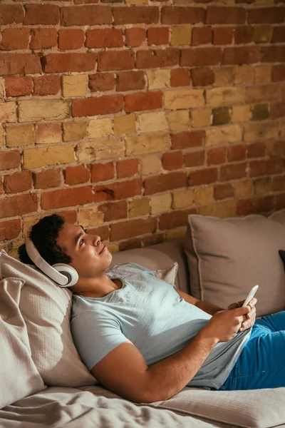 Schöner Mann mit zwei Rassen, der während der Isolation mit Kopfhörern und Smartphone auf dem Sofa chillt und Musik hört — Stockfoto