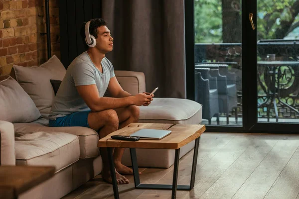 Giovane uomo di razza mista agghiacciante e ascoltare musica con cuffie e smartphone sul divano durante l'isolamento — Foto stock