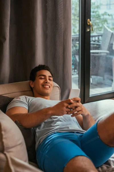 Молодий щасливий змішаний гоночний чоловік охолоджує і використовує смартфон на дивані під час карантину — стокове фото