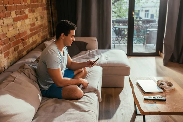 Smiling mixed race man chilling and using smartphone on sofa in living room with gadgets during quarantine — Stock Photo