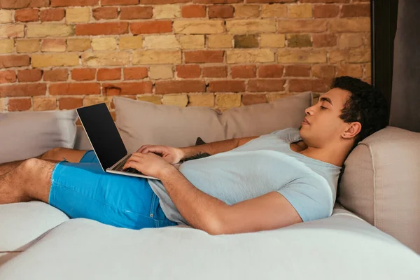 Somnoliento hombre de raza mixta usando portátil con pantalla en blanco en el sofá en casa en cuarentena - foto de stock