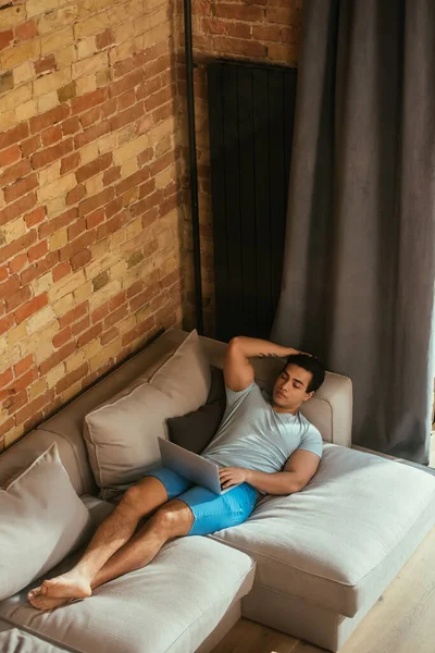 Молодий змішаний гоночний чоловік охолоджує ноутбук на дивані під час самоізоляції — стокове фото