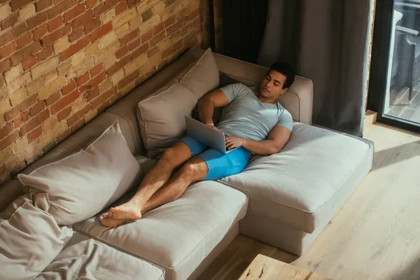 Homme de race mixte utilisant un ordinateur portable et se refroidissant sur le canapé à la maison en quarantaine — Photo de stock