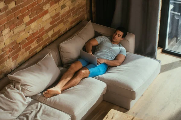 Hombre de raza mixta utilizando el ordenador portátil y escalofriante en el sofá en casa en cuarentena - foto de stock