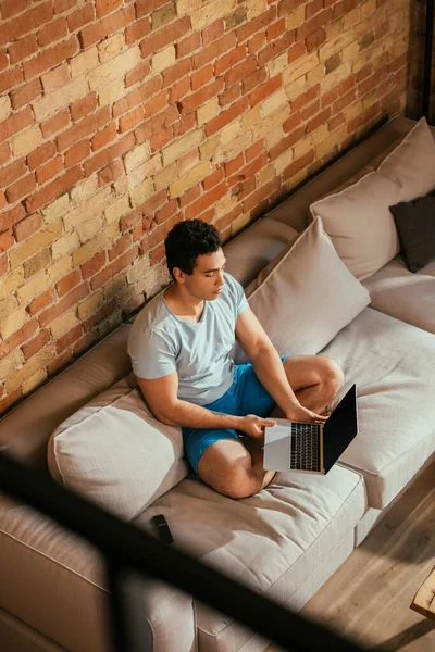 Hombre bi-racial que sostiene el ordenador portátil con la pantalla en blanco en el sofá en casa en cuarentena - foto de stock