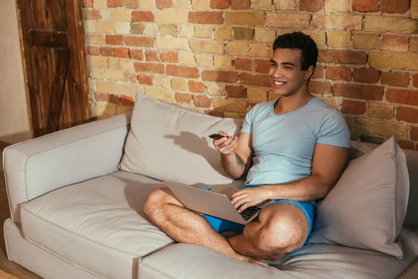 Sorrindo homem de raça mista com laptop segurando controlador remoto e assistindo tv na sala de estar durante a quarentena — Fotografia de Stock