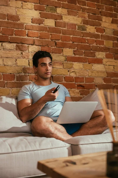 Mixed race man with laptop holding remote controller and watching tv in living room during self isolation — Stock Photo