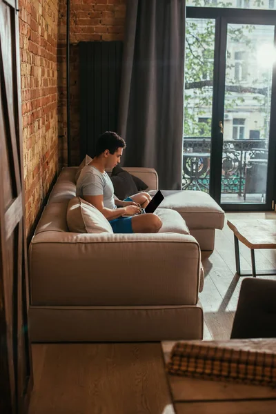 Homme de race mixte utilisant un ordinateur portable avec écran blanc sur le canapé à la maison en quarantaine — Photo de stock